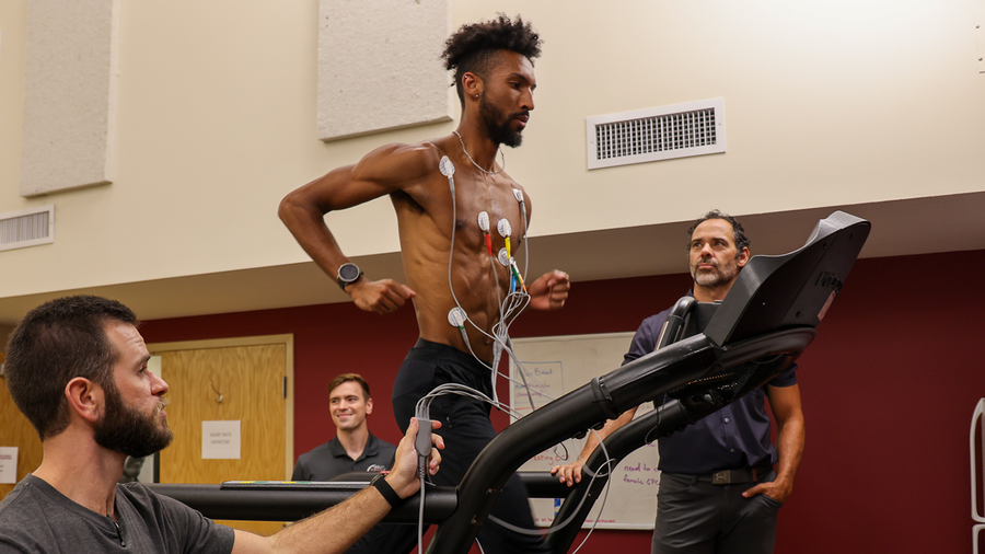 A man runs on a treadmill with electrodes attached to his chest while three people monitor him and the equipment
