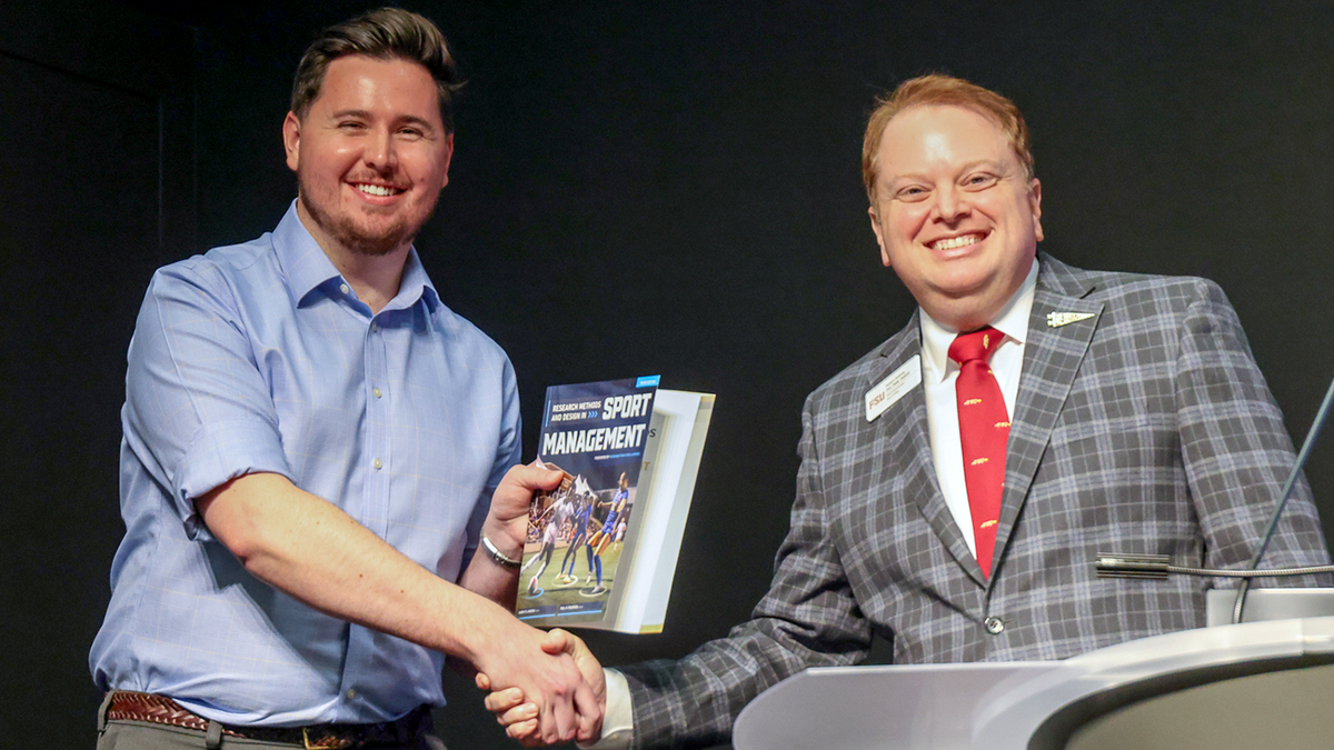 Graphic Designer Colton Flynn holds a textbook while shaking hands with Anne's College Dean Damon Andrew